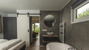 A modern bedroom with a large bed, a walk-in closet, a bathroom with a freestanding bathtub, a round mirror, and a window. - Cottage with Scenic Pasture Outlook (Zuna)