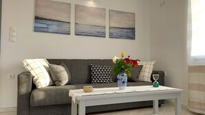 Sala de estar com sofá cinza, mesa de centro branca e um vaso com flores. Há três quadros na parede e uma janela com cortinas.