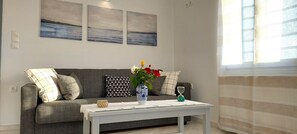 A living room with a grey sofa, a white coffee table, and a vase with flowers. There are three paintings on the wall and a window with curtains. - Spacious apartment with sea view (Paros)