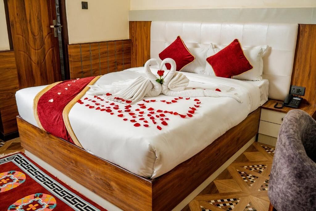 Cama bem arrumada com arranjo de pétalas de rosa em formato de coração e toalha em forma de cisne, travesseiros vermelhos e cabeceira de madeira.
