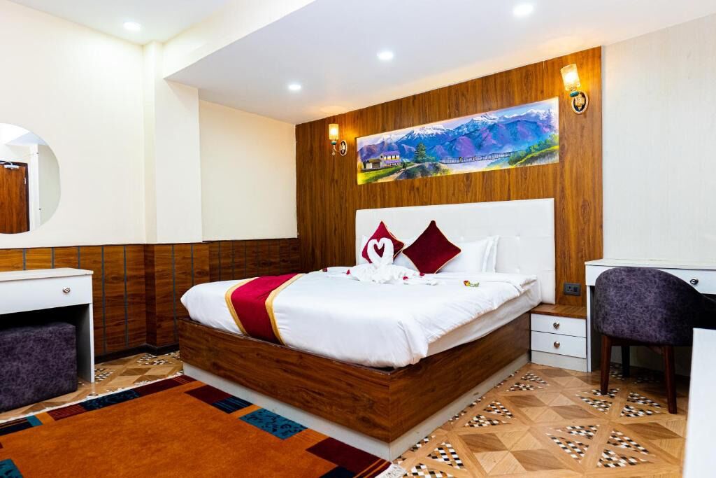 Habitación de hotel con cabecera de madera, cama con ropa de cama blanca y detalles rojos, un escritorio con una silla y un cuadro en la pared.