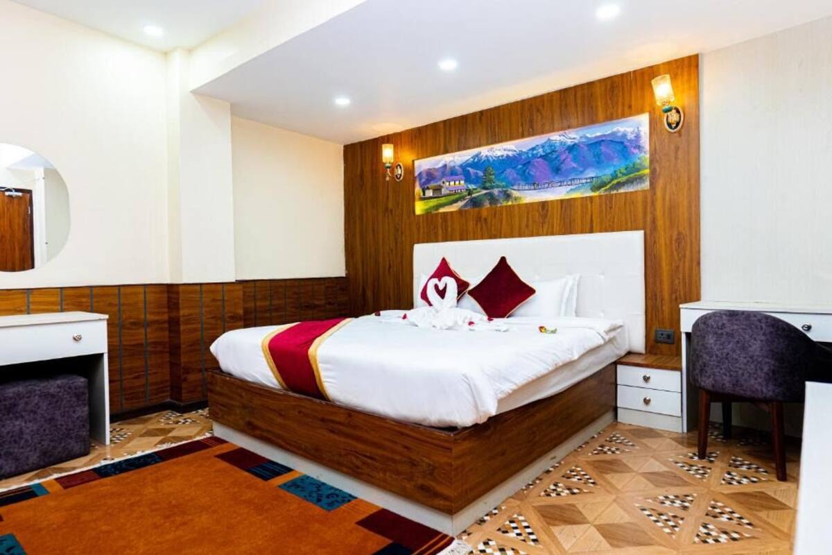 A hotel room with a wooden headboard, a bed with white bedding and red accents, a desk with a chair, and a painting on the wall.