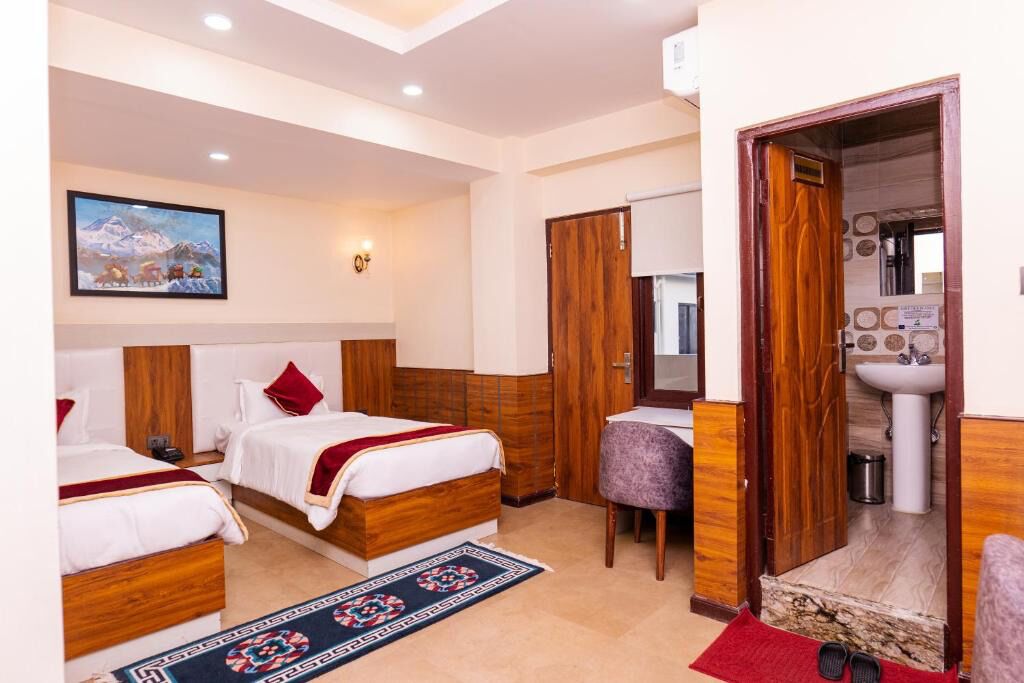 A hotel room with three beds, a small table, a chair, a bathroom with a sink, and a painting on the wall.