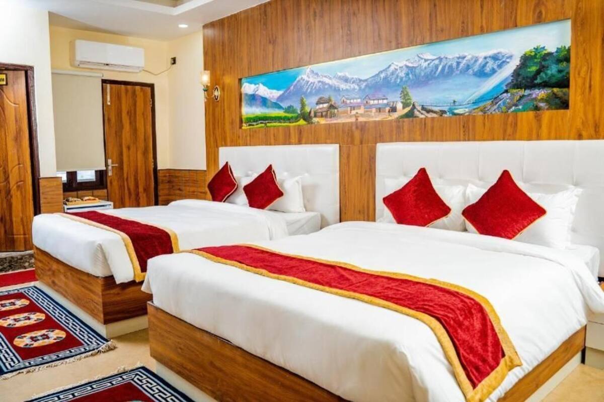 A hotel room with two beds, wooden headboards, a painting of a mountainous landscape, and a red and white patterned rug.