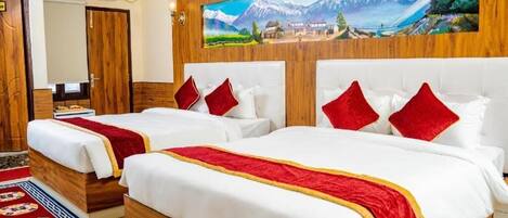A hotel room with two beds, wooden headboards, a painting of a mountainous landscape, and a red and white patterned rug.