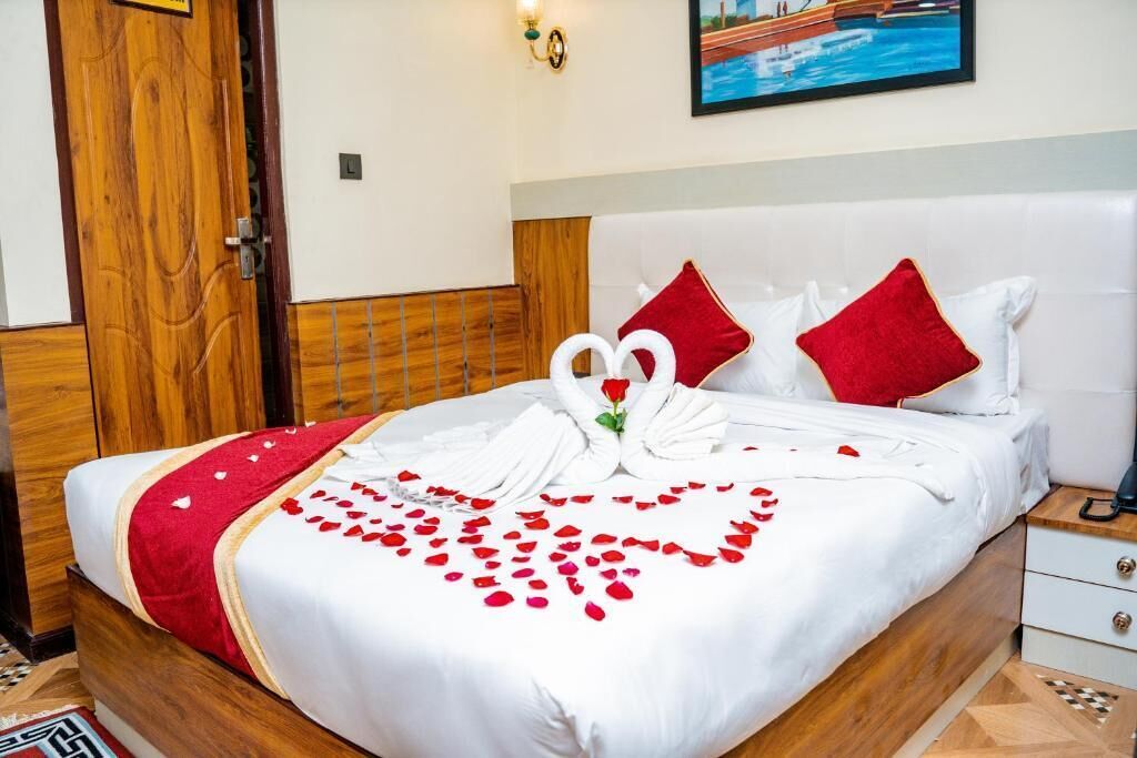 A hotel room with a neatly made bed, a wooden headboard, a white nightstand, and a wall-mounted light fixture.