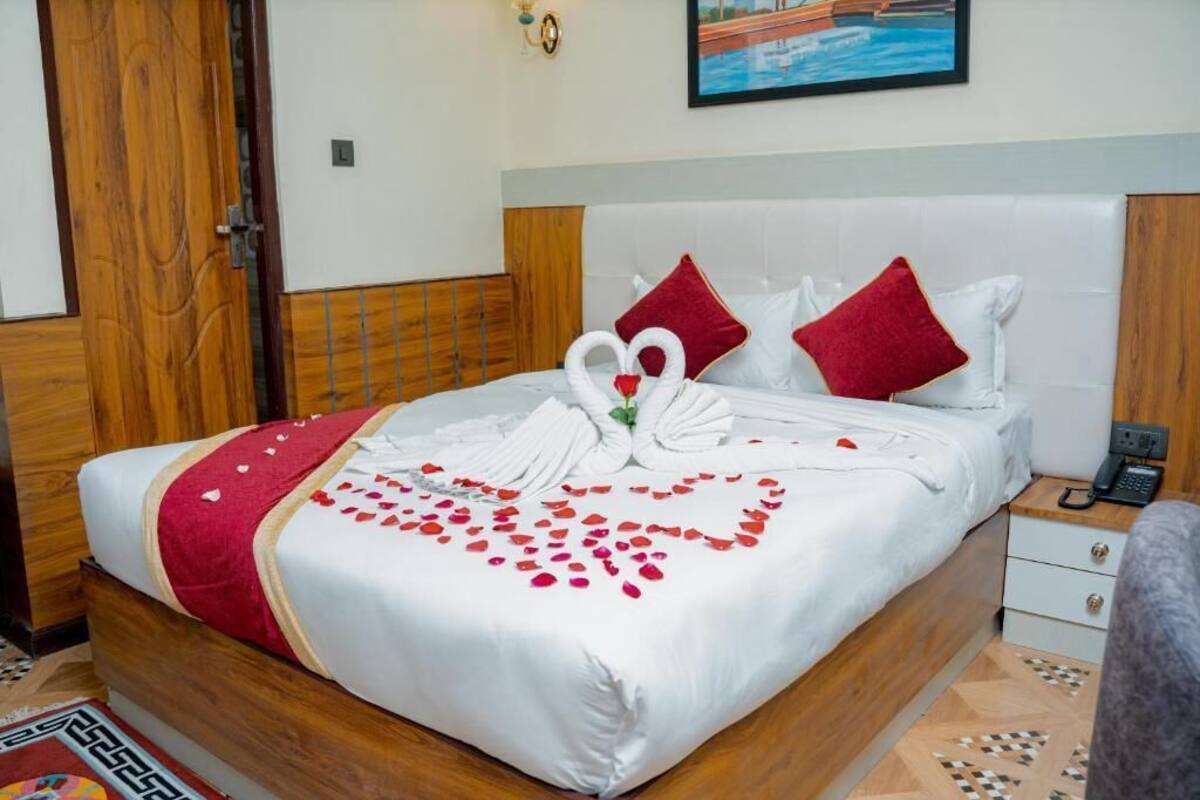 A neatly made bed with a heart-shaped arrangement of rose petals and white towels, a wooden door, a white bedside table with a phone, and a painting on the wall.