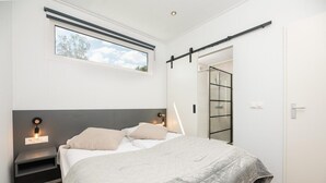 Une chambre à coucher comprenant un lit, des tables de chevet, une fenêtre, une porte coulissante et un luminaire fixé au mur.