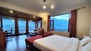 Una habitación de hotel con una cama grande, un área de estar con una mesa y vistas a montañas a través de amplios ventanales.