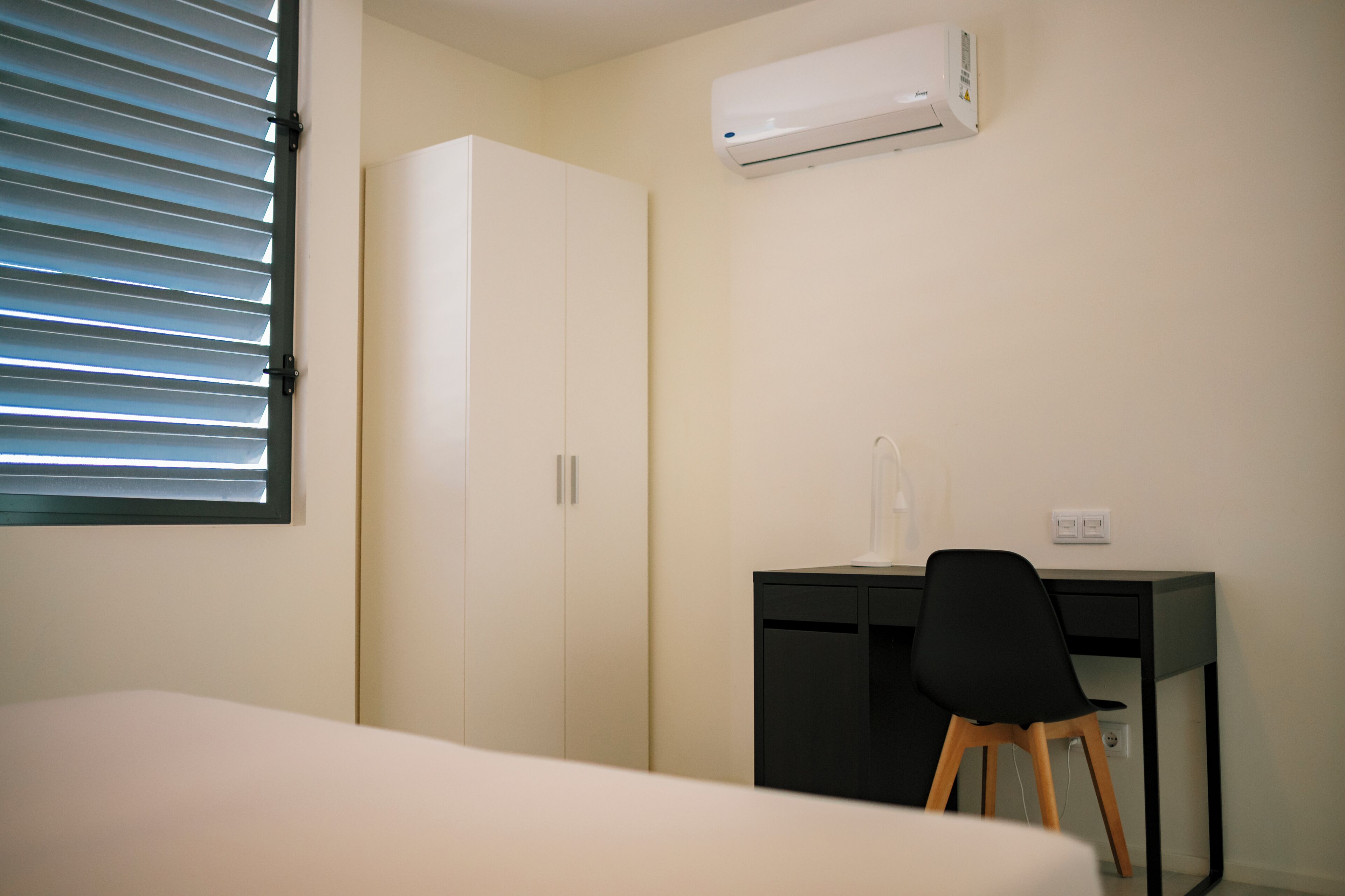 Ein Schlafzimmer mit Bett, Schreibtisch mit Stuhl, Kleiderschrank und Klimaanlage.