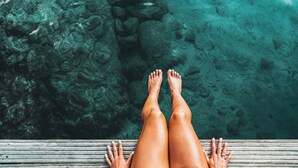 Un muelle de madera con las piernas y las manos de una persona, con vista a aguas claras donde se ven rocas y vida acuática.