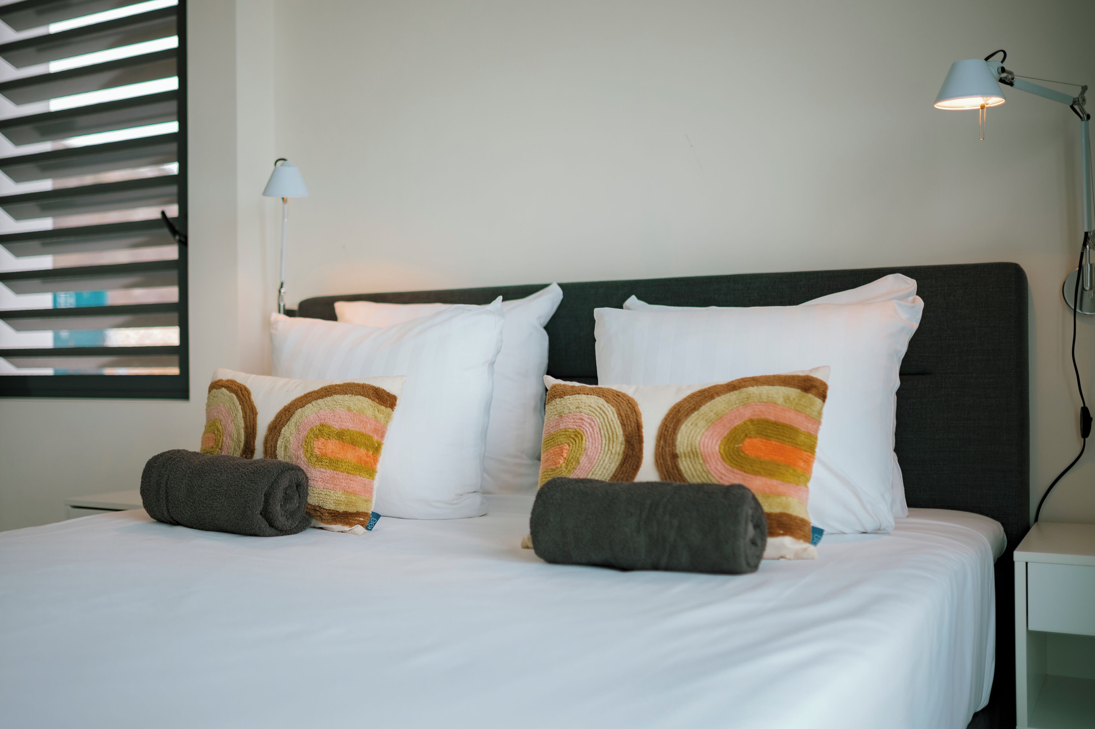 A neatly made bed with white and colorful pillows, a dark headboard, and a lamp on a bedside table.