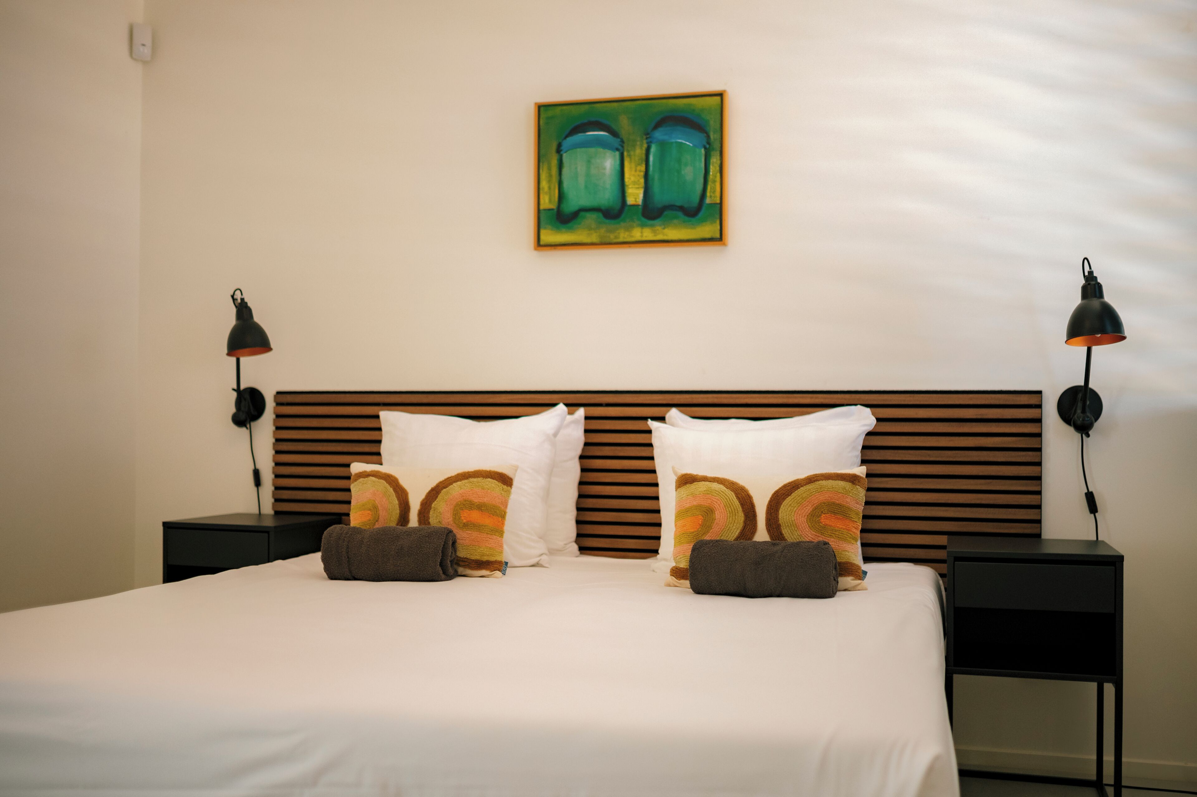 Una cama bien tendida con ropa de cama blanca, dos mesitas de noche negras con lámparas, y un cuadro abstracto de colores sobre el cabecero.