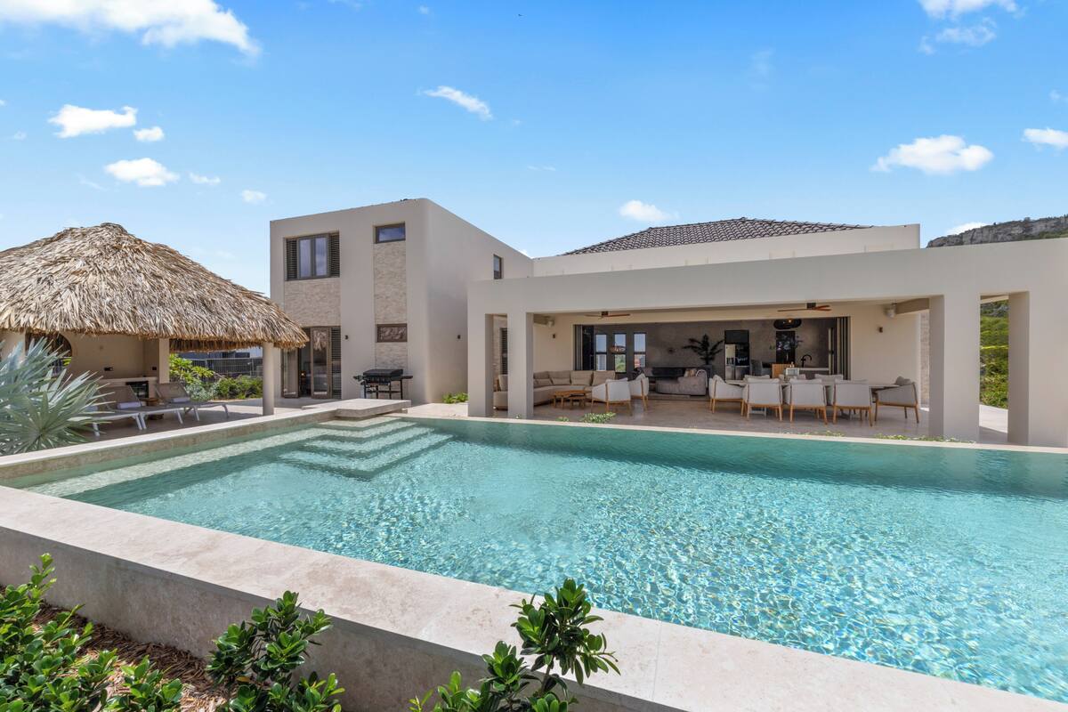 Una casa moderna con piscina, mobiliario de exterior y una estructura con techo de paja.
