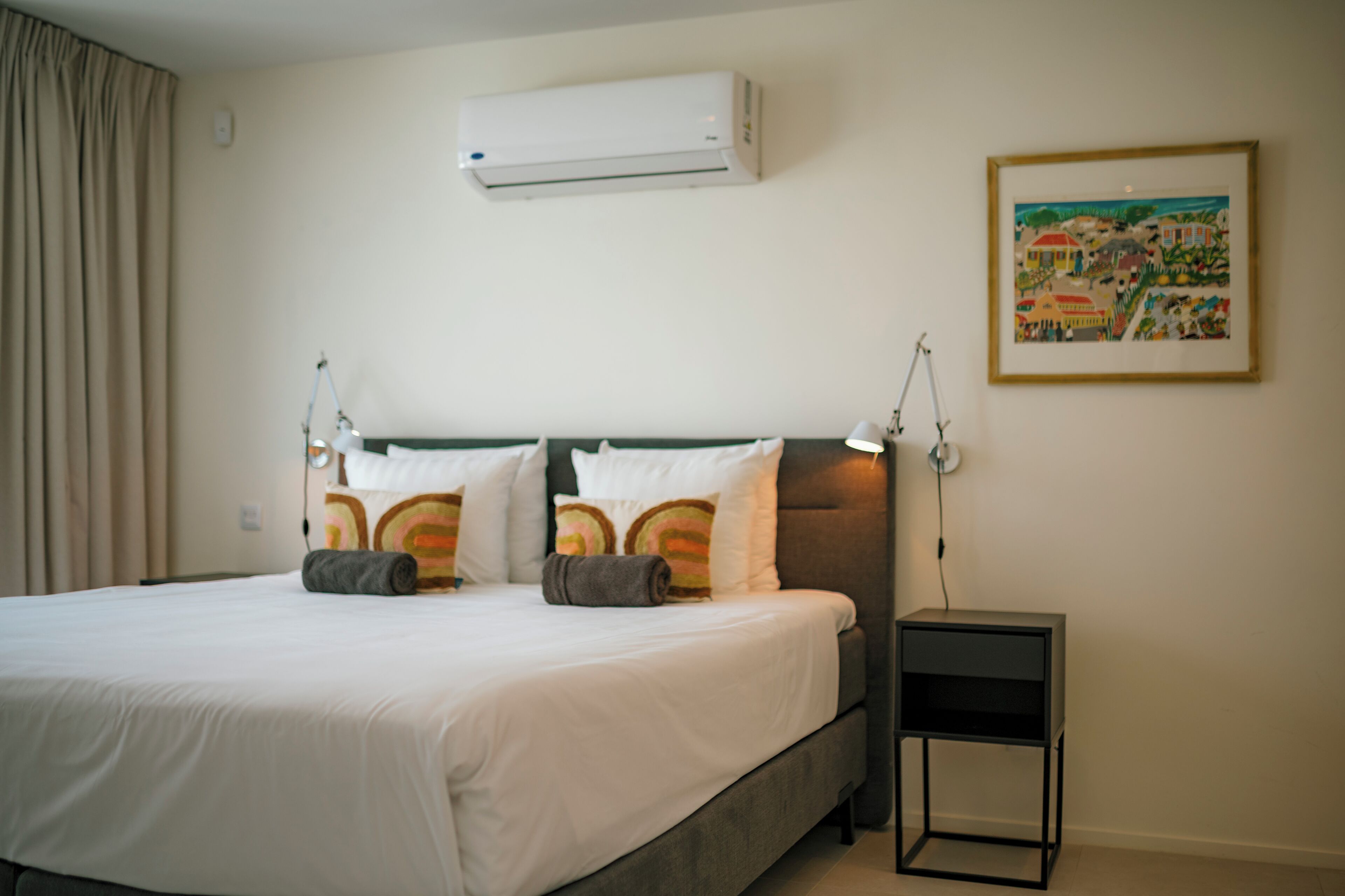 A hotel room with a bed, an air conditioner, a nightstand, and a painting on the wall.