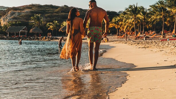 Escena de playa con una pareja caminando por la orilla, palmeras y sombrillas.