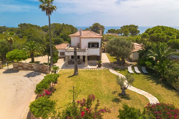 A two-story house with a tiled roof, surrounded by a well-maintained garden with palm trees and flowering plants, and a view of the sea.