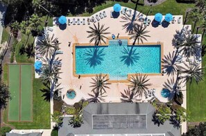 Eine Vogelperspektive zeigt ein Resort mit einem zentralen Schwimmbecken, Liegestühlen, Palmen und einem Tennisplatz.
