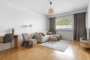 Ein modernes Wohnzimmer mit Holzboden, einer Couch, einem Sessel, einem kleinen Tisch, einem Fernseher und einem Fenster mit Vorhängen.