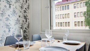 Una mesa de comedor dispuesta con platos, vasos y cubiertos, una silla y un ventanal con vista a un edificio.