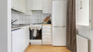 Kompakti keittiö, jossa on valkoiset kaapit, sisäänrakennettu uuni, jääkaappi ja ikkuna, jossa on verho.