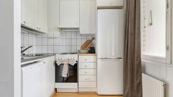A compact kitchen with white cabinets, a built-in oven, a refrigerator, and a window with a curtain.