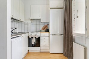 Eine kleine Küche mit weißen Schränken, einem Einbauofen, einem Kühlschrank und einem Fenster mit Vorhang.
