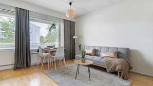 Moderni olohuone, jossa on harmaa sohva, pyöreä sohvapöytä, ruokapöytä tuoleineen ja amppelissa roikkuva kasvi.