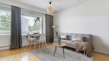 Moderni olohuone, jossa on harmaa sohva, pyöreä sohvapöytä, ruokapöytä tuoleineen ja amppelissa roikkuva kasvi.