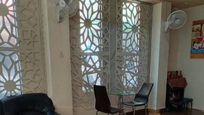A room with a checkered floor, a black leather sofa, a glass table with two chairs, decorative screens, and two standing fans. - Del Mar Hotel (Pattaya)