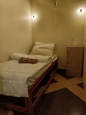 A small, dimly lit room with a single bed, a wooden cabinet, and a small table with a bottle and glasses. - Del Mar Hotel (Pattaya)