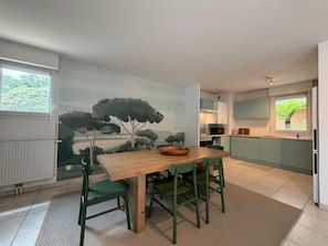 A dining area with a wooden table and green chairs, a kitchen with teal cabinets, and a mural of trees on the wall. - L'Escale de Brindos - Collective pool (ANGLET)