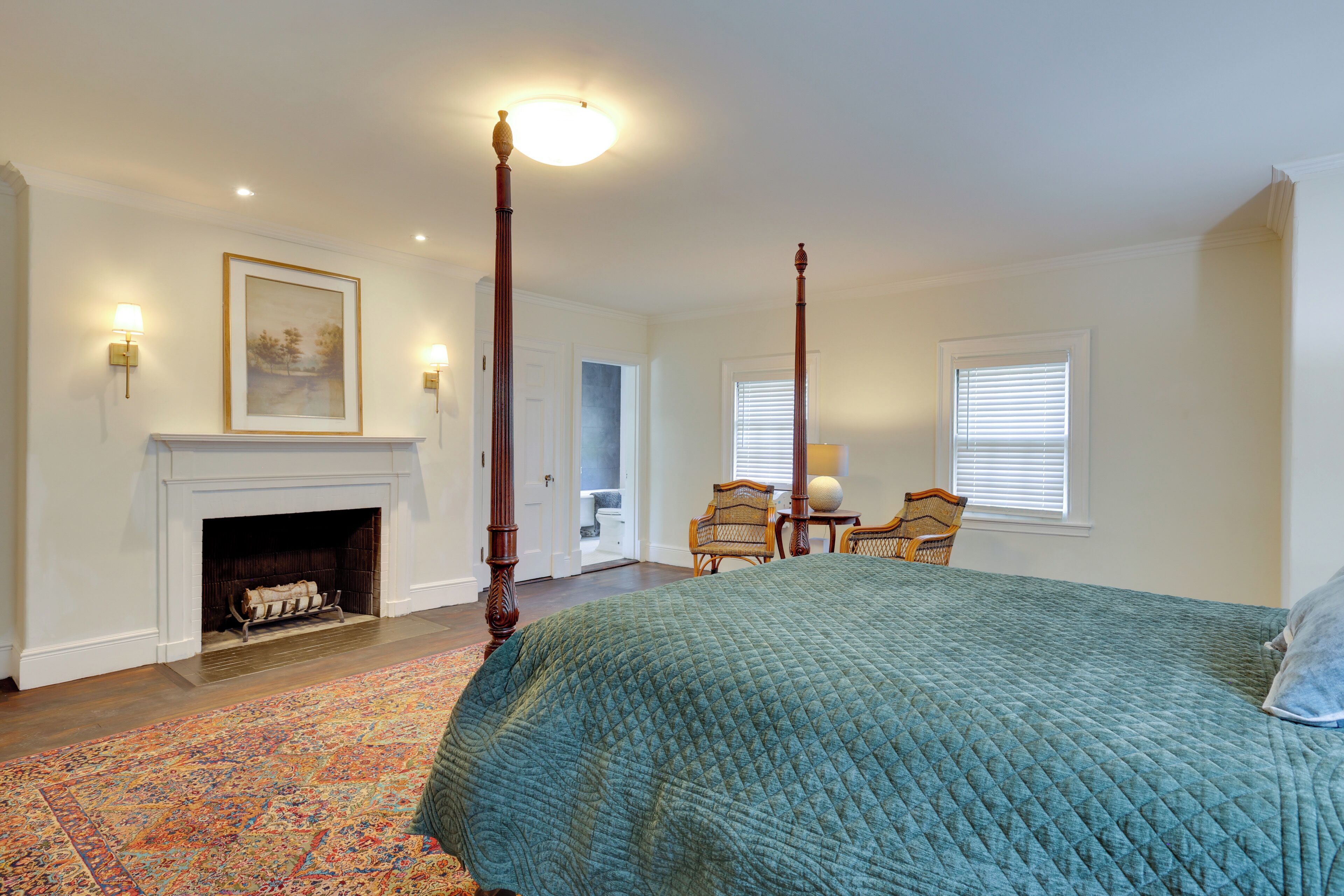 A bedroom with a large bed, a fireplace, a door leading to another room, and a window with blinds.