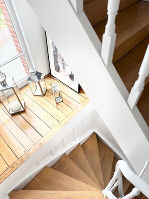 Un escalier en bois avec des rampes blanches, une fenêtre donnant sur un mur de briques et une petite table sur laquelle se trouvent une bougie et un cadre photo.