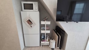 A compact kitchenette with a microwave, a refrigerator, a white shelf unit, and a black radiator.