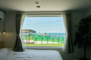 A hotel room with a large window, a bed, curtains, a plant, and a view of the beach and ocean.