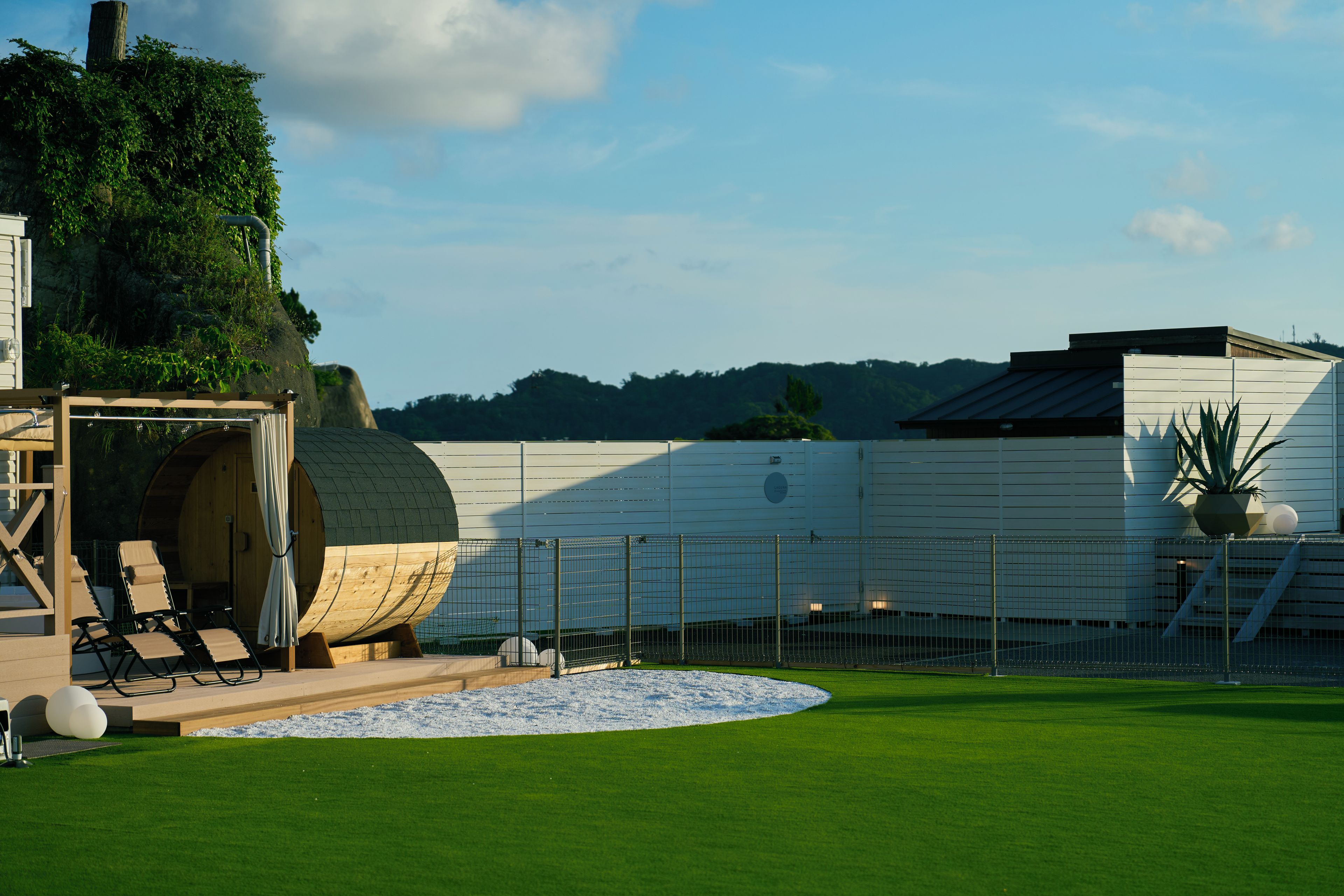 A well-maintained lawn with a wooden deck and lounge chairs, a large white building with a flat roof, and a fenced area with a circular white space.