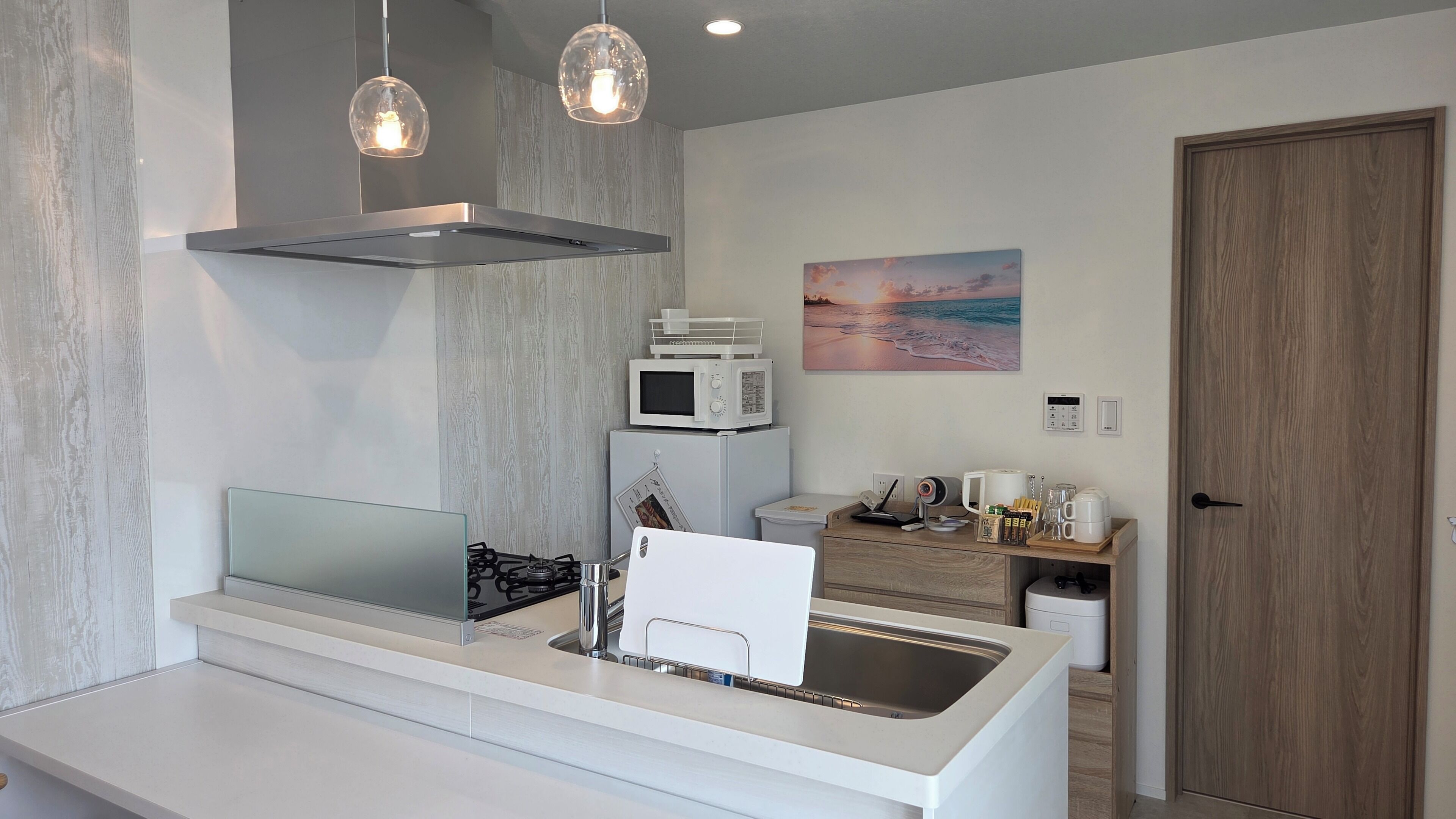 Una cocina moderna con estufa a gas, microondas y pileta. Hay luces colgantes, un cuadro en la pared y una puerta de madera.