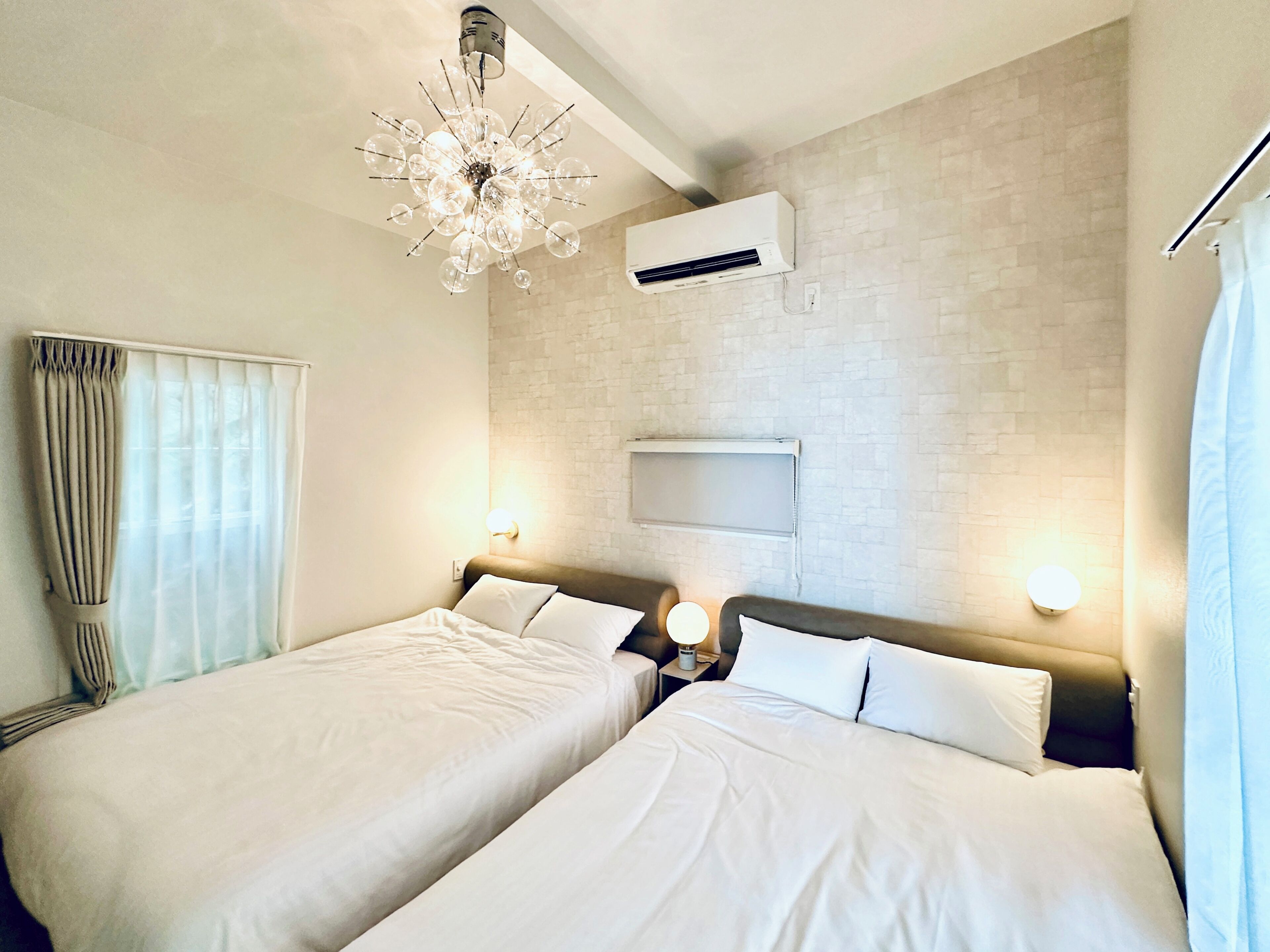 Een hotelkamer met twee bedden, een moderne kroonluchter, een airconditioner en een bakstenen muur.