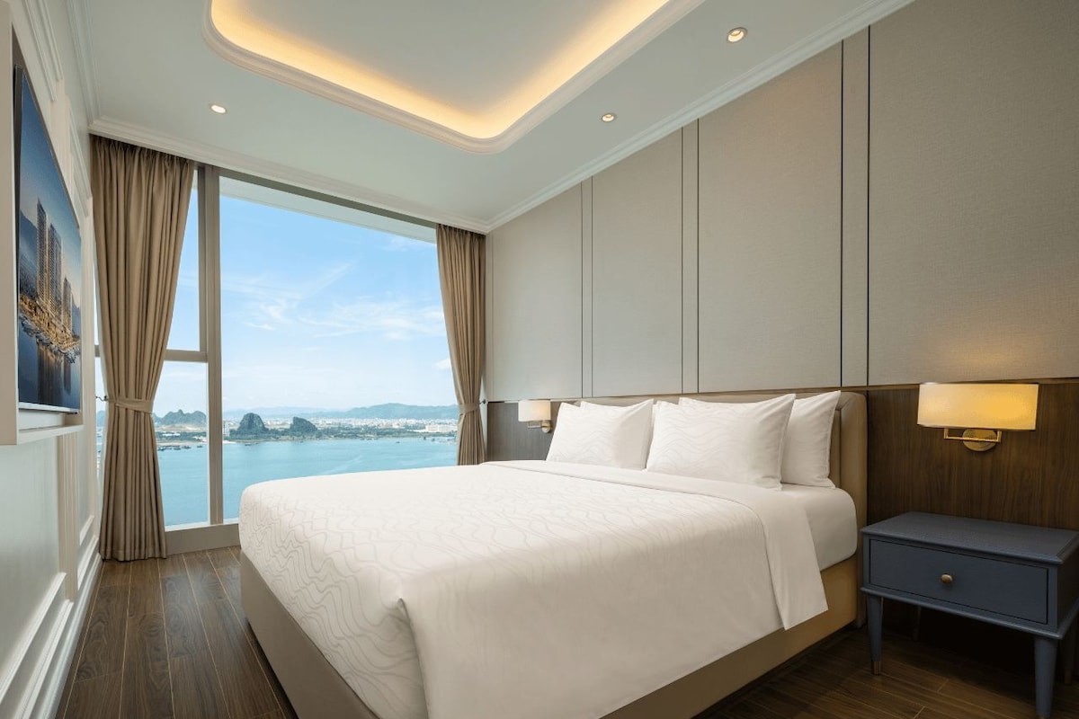 A hotel room with a large bed, a nightstand, a wall-mounted lamp, and a view of the sea.