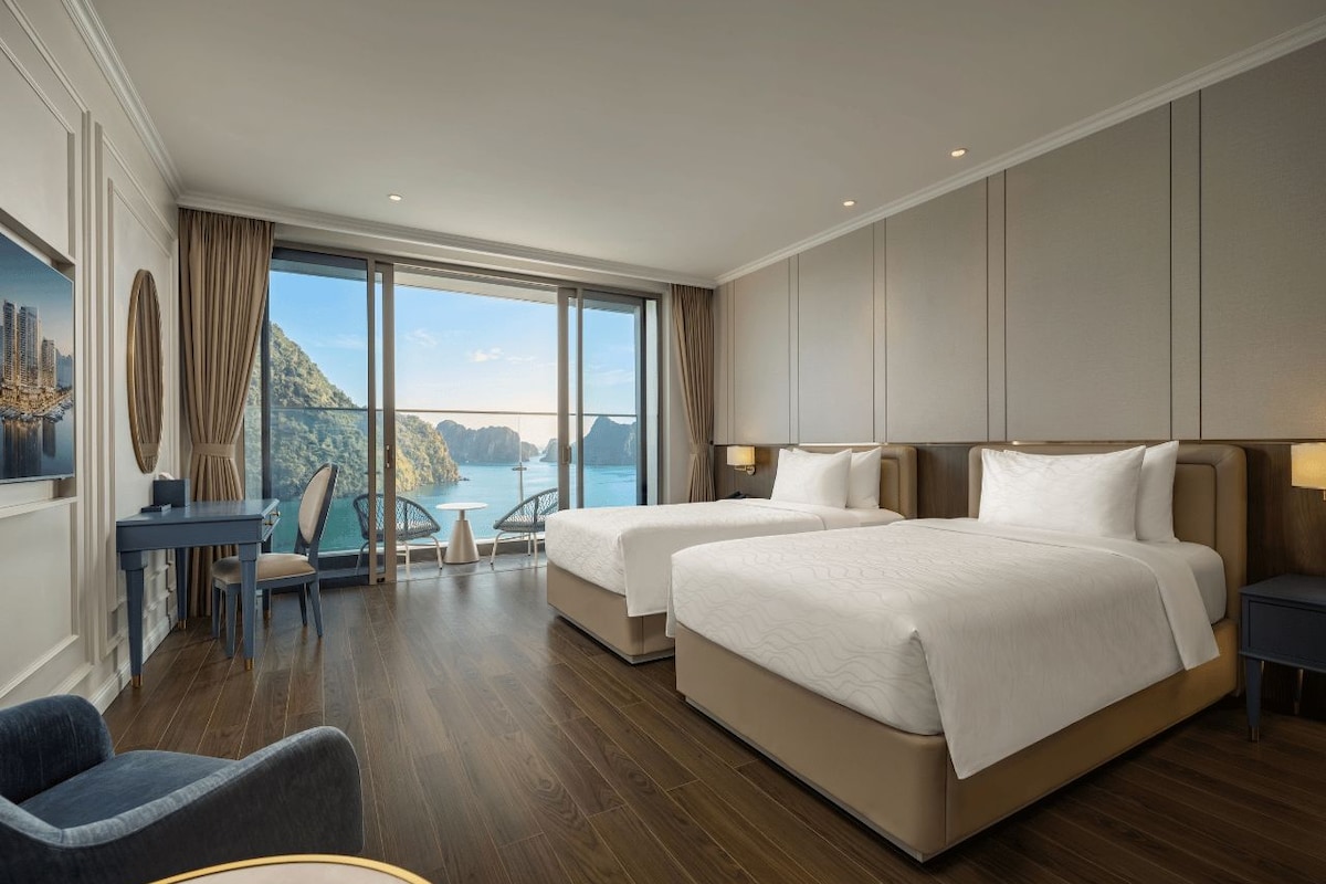 A hotel room with two beds, a desk, a chair, and a view of the sea.