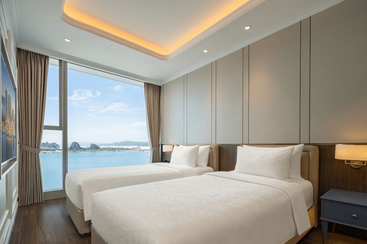 A hotel room with two beds, a view of the sea, and a nightstand with a lamp.