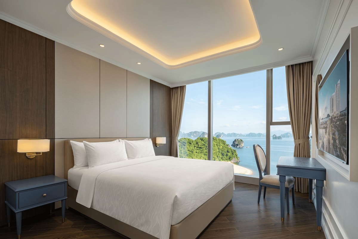 A modern hotel room with a large bed, a desk, and a view of a coastal landscape.
