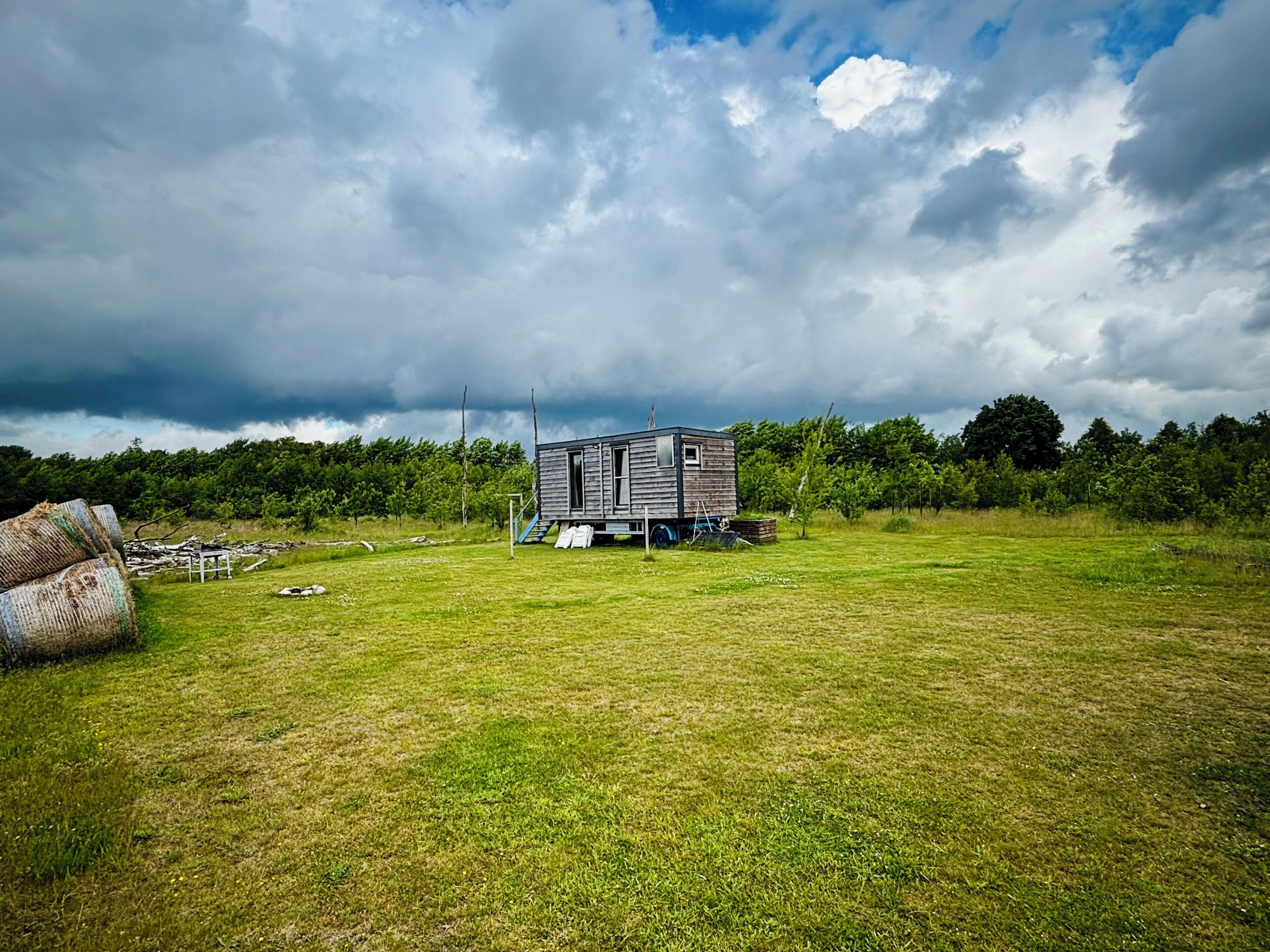 A small, single-story cabin with a porch, surrounded by a grassy field and trees, under a cloudy sky.
