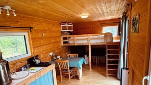 A cabin interior with a kitchen area, a dining table with chairs, a bunk bed, and a window providing a view of the outdoors. - Tinyhouse idyll: nature & chickens (Aurich)