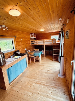 A cabin interior with a kitchen area, a dining table with chairs, a bunk bed, and a window providing a view of the outdoors. - Tinyhouse idyll: nature & chickens (Aurich)