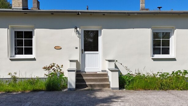 Ein zweigeschossiges Haus mit weißer Fassade, einer Haustür, zwei Fenstern und einer kleinen Veranda mit Stufen zum Eingang.