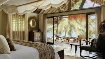 A bedroom with a bed, a nightstand, a mirror, and a view of the beach through large windows.