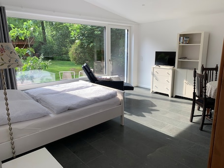 A modern bedroom with a bed, a TV, a bookshelf, and a view of a garden.. Vacation apartment Heske, 37 m², 1-2 pers.