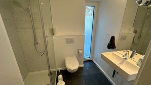 Une salle de bain moderne dotée d’une douche à parois de verre, de toilettes, d’un lavabo avec un miroir et d’une fenêtre laissant entrer la lumière naturelle.